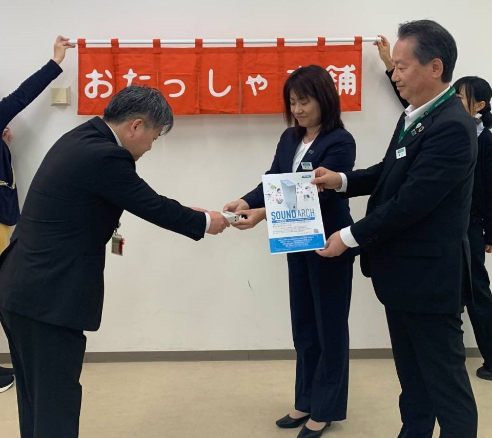 「おたっしゃ本舗」と書かれた赤い暖簾の前で、スーツ姿の井上典子さんと尾上兼一さんが並んで立ち、山崎副部長が頭を下げパンフレットと小型機器を受け取っているる様子の写真