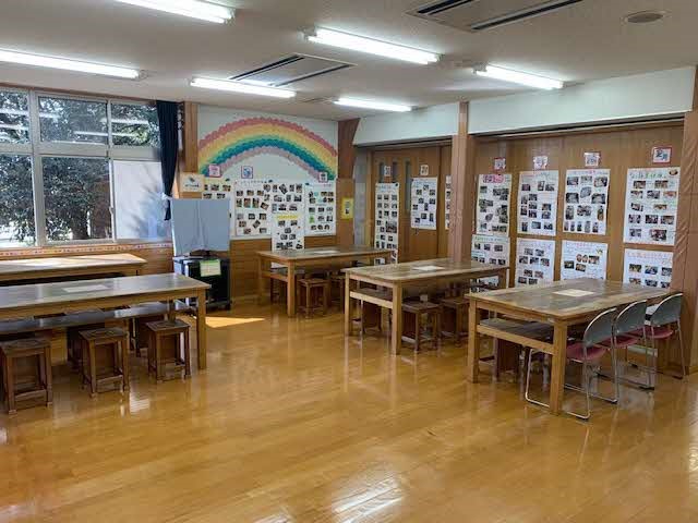 壁一面に写真掲示と虹の装飾がある広々とした明るい教室で、複数の木製机と椅子が整然と配置された写真