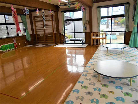 大きな窓のある明るい室内の天井に飾られたこいのぼりが揺れ、象柄マット上の円卓と木床の広々とした遊びスペースが広がる写真