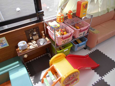 色付きジョイントマット上に小型滑り台とおままごと用玩具棚が並び、隣にソファが置かれた親子ふれあいルームの写真