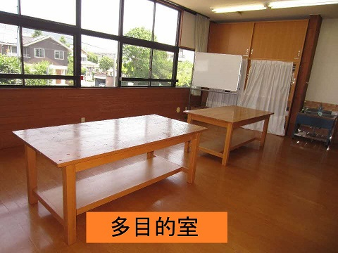 大きな窓から光が入る明るい室内に木製の作業台とホワイトボードが配置された多目的室の写真
