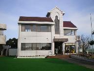 白い外壁と赤い屋根を持つ二階建ての川副児童館が青空の下で正面入口と芝生を前にして立つ写真