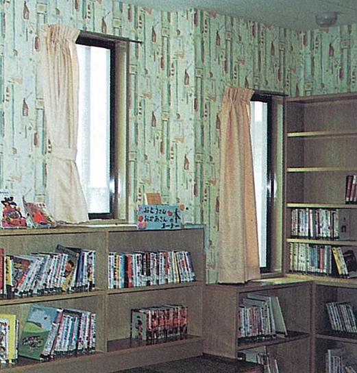窓際に淡色カーテンと模様壁紙があり、低い本棚に多数の書籍が並ぶ落ち着いた図書室の写真