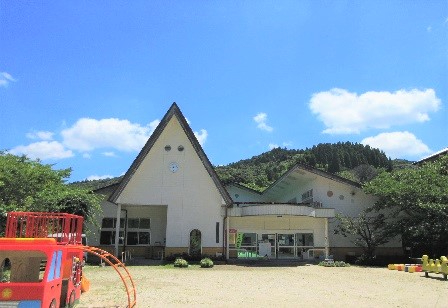 青空の下で三角形の大きな屋根が特徴的な白い園舎が建ち、手前の砂場のような広場には赤いバスを模した遊具やオレンジ色のトンネル型遊具が設置された松梅保育所の写真
