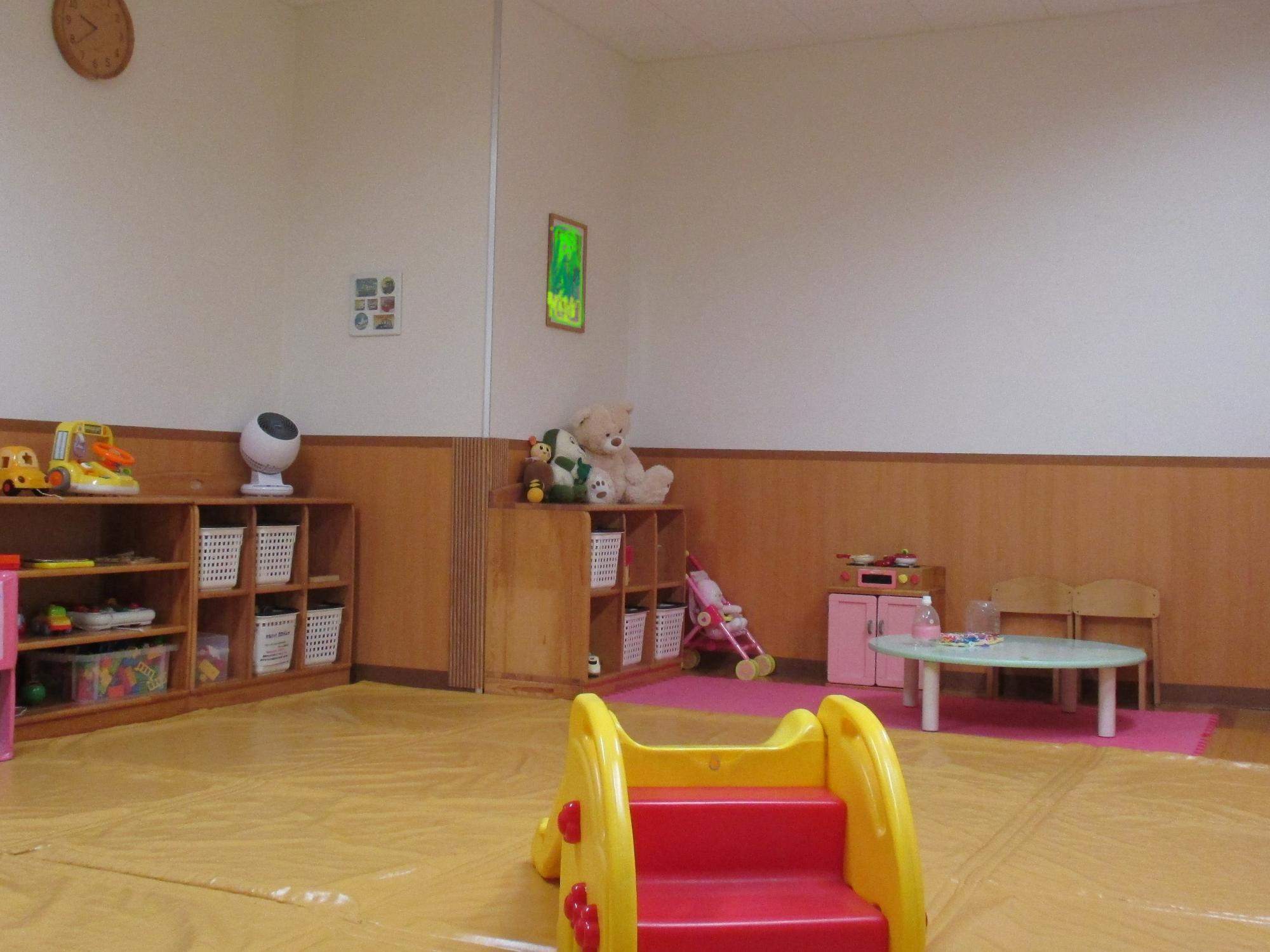 木の棚におもちゃやぬいぐるみが並ぶ広い室内に小さな滑り台や低い机と椅子が整えられた明るい保育室の写真