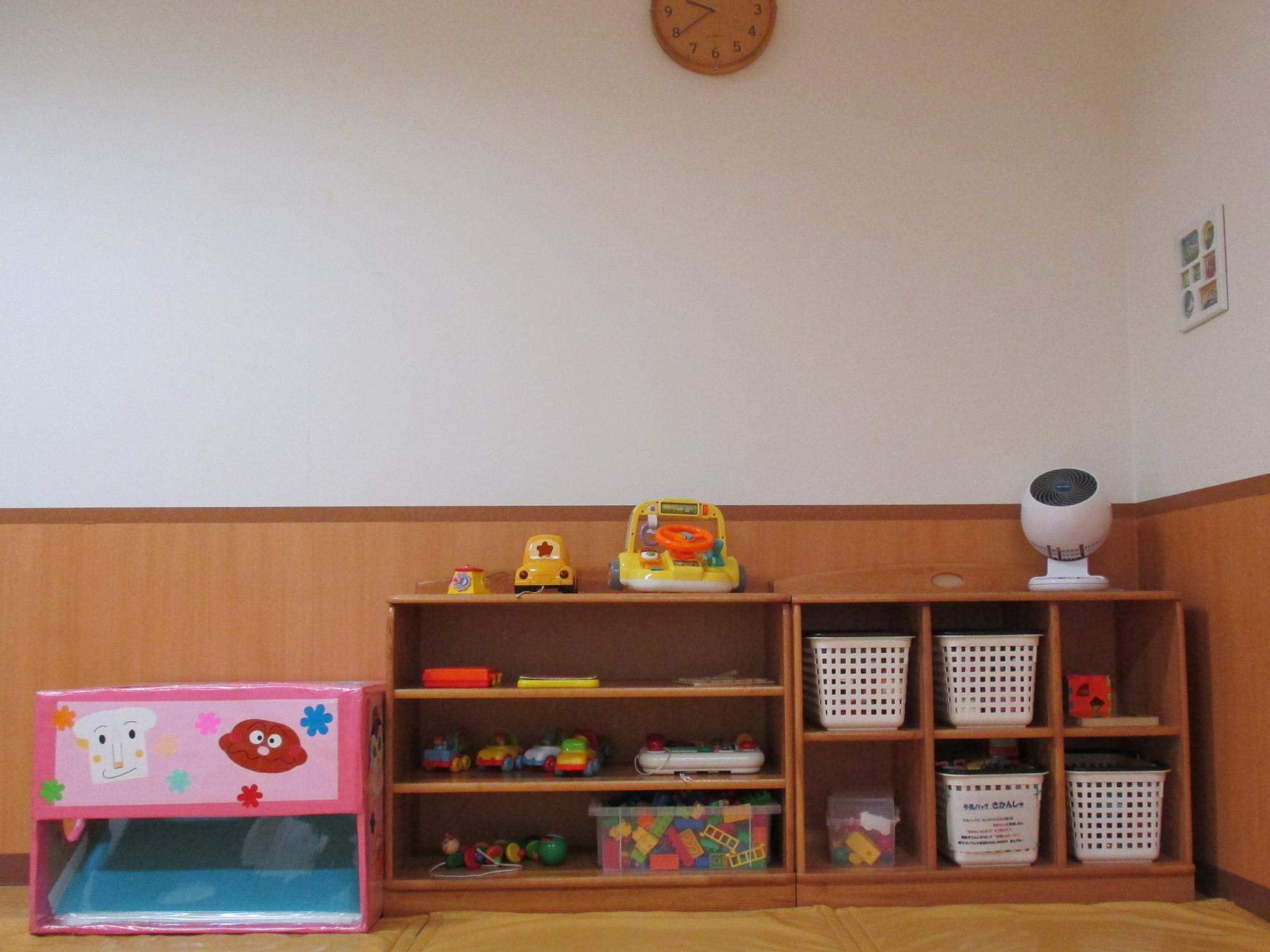 木製棚に積み木や乗り物のおもちゃ、白いバスケットが整然と並び壁掛け時計が見える保育室の写真