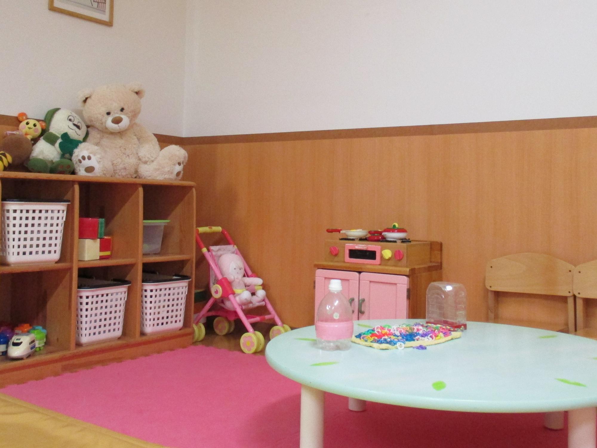 棚に並ぶぬいぐるみやままごとセット、低い丸机と椅子が置かれた明るい保育室の一角を捉えた写真