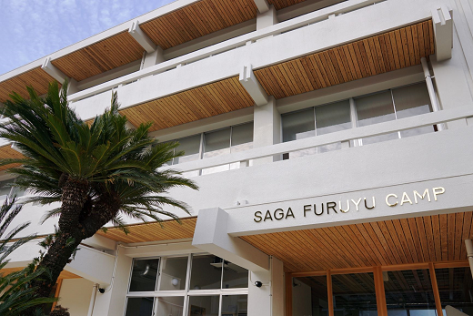 白を基調としたモダンな建物のエントランスに「SAGA FURUYU CAMP」という金色の文字が掲げられ、木材を多用した軒天と南国風のヤシの木が植えられた佐賀古湯キャンプの外観写真