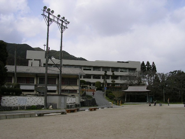 山々や2階建ての古い建物と、その背後に近代的な白い数階建ての大きな建物を背景に、奥に照明塔がそびえ立つ広い土のグラウンドの写真
