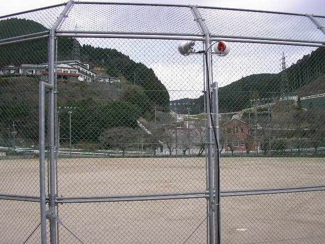 山々に囲まれた屋外運動場において、バックネットの金網越しに土のグラウンドが広がっている写真