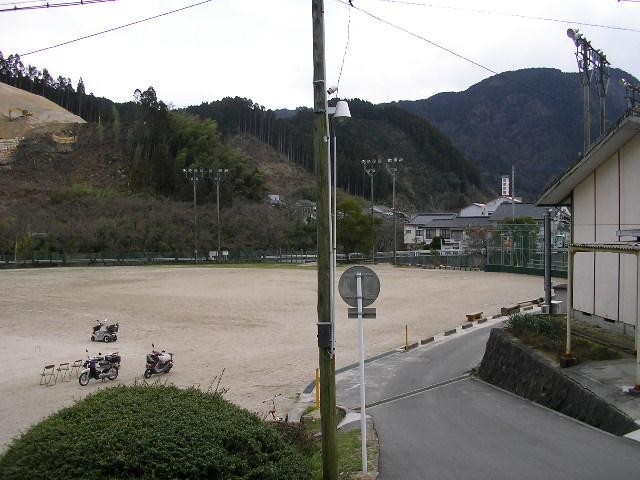 山々に囲まれた平坦で広い土のグラウンドの隅に、数台のバイクや折りたたみ椅子が置かれ、手前の坂道や電柱越しに静かな集落の建物や緑豊かな斜面が広がる、屋外運動場の写真