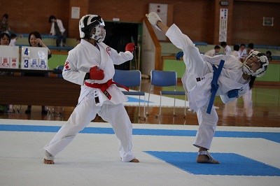 赤帯と青帯の空手選手が試合場で構え合い、青帯の選手が高い回し蹴りを放ち赤帯の選手が受け止めようとする迫力ある瞬間を捉えた写真