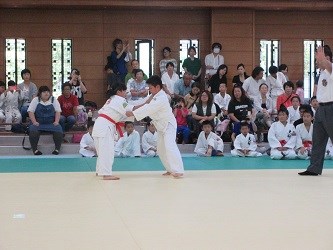 畳の上で二人の柔道選手が組み合って体勢を探りながら技を仕掛ける瞬間を、多くの観客が見守る試合風景の写真