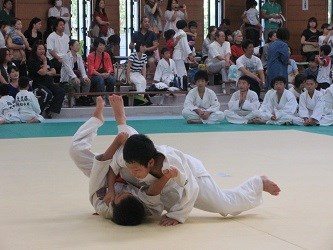 畳の上で二人の柔道選手が組み合いながら技を掛け合い、周囲の選手や観客が真剣に見守る試合の緊迫した瞬間を捉えた写真