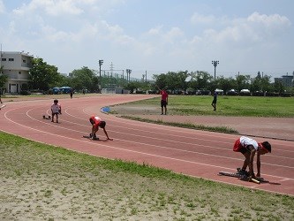 陸上トラックで子どもたちがスターティングブロックに構えて号令を待ち、青空の下で緊張感が漂う様子を捉えた写真