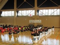 体育館の中央で複数のスポーツチームが背番号入りのユニフォーム姿で整然と座り、木床が光る広い空間の中、掲示された横断幕や国旗の前で開会式の開始を静かに待つ様子を収めた写真