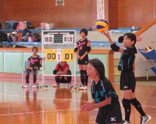 体育館で黒のユニフォームの選手が力強くサーブしようとしている瞬間を、仲間が集中して見守る緊張感ある試合風景の写真