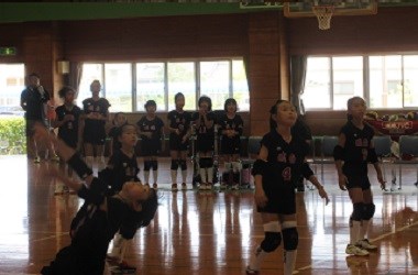 体育館で黒いユニフォームの選手たちがボールを追って連携しながらプレーし、背後では仲間が緊張感をもって見守る写真