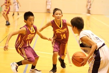 体育館で白のユニフォームの選手がドリブルから攻め込もうとし、紫のユニフォームの選手二人が素早く挟み込んで守備する様子を捉えたバスケットボールの試合写真