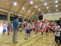 体育館で子どもたちが旗を高く掲げ整列し、一人の選手が指導者の前で選手宣誓している写真
