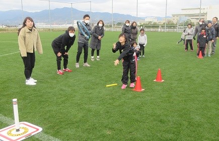 芝生の広場で子どもがリングを的へ勢いよく投げ、周囲の仲間や大人が見守る中で競技が進む様子を捉えた写真
