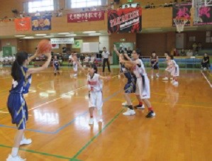 体育館で青いユニフォームの選手がパスを狙い、白い選手たちが守備につき緊迫した攻防が展開されるバスケットボールの試合写真