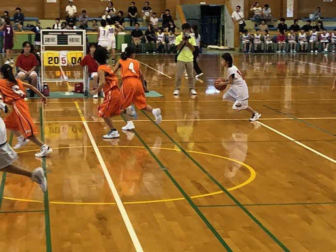 体育館のコートで白いユニフォームの選手がドリブルし、橙色の選手数名が素早く囲みながら攻防が展開されている様子を捉えたバスケットボールの試合写真