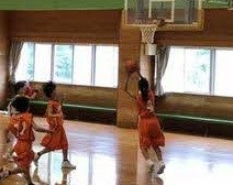 体育館で橙色のユニフォームの選手がリング下でシュートを放ち、後方から仲間が走り込む瞬間を捉えた写真