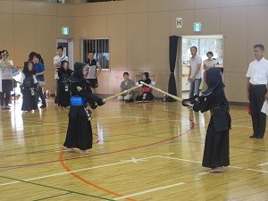 体育館で防具を着けた二人の剣道選手が竹刀を交えて向かい合い、周囲で観客が静かに見守る様子を捉えた写真