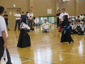 体育館で防具を着けた二人の剣道選手が竹刀を交えて攻防を繰り広げ、周囲で審判や観客が熱心に見守る様子を捉えた写真