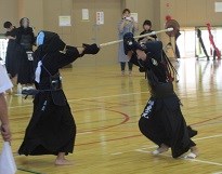 体育館で防具を着けた二人の剣道選手が前傾姿勢で竹刀を交えて激しく攻防し、周囲の観客が静かに見守る写真