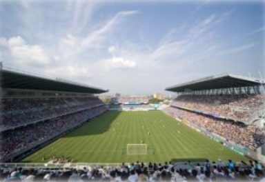 満員の観客で埋まった大型サッカースタジアムを上方から眺め、青空の下に広がる緑のピッチと観客席全体を捉えた写真