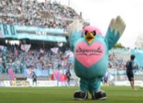 ピンクの胸元が特徴の青い鳥型マスコットのウィントスが賑わう観客席を背に両翼を大きく広げてピッチ上に立つ姿を捉えた写真