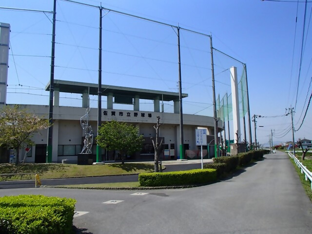 快晴の青空の下で「佐賀市立野球場」の文字が掲げられた球場の正面外観が写っており、高い防球ネットの支柱が並び立ち、手前には舗装された道路と整えられた植栽、そして銀色のオブジェが配置されている、野球場正面の写真