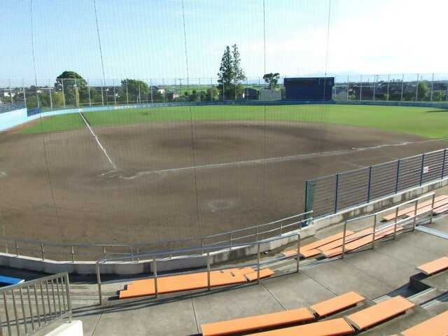 佐賀市立野球場の観客席から見下ろした内野グラウンドの全景で、手前にはオレンジ色の座席が並ぶスタンドがあり、防球ネット越しに整備された土のダイヤモンドと緑の芝生の外野、そして遠くの青空と街並みが広がる、球場内の写真