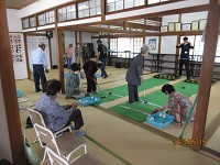 畳敷きの広間で高齢者たちが緑のマット上に置かれた用具を使って楽しげに競技を行う写真