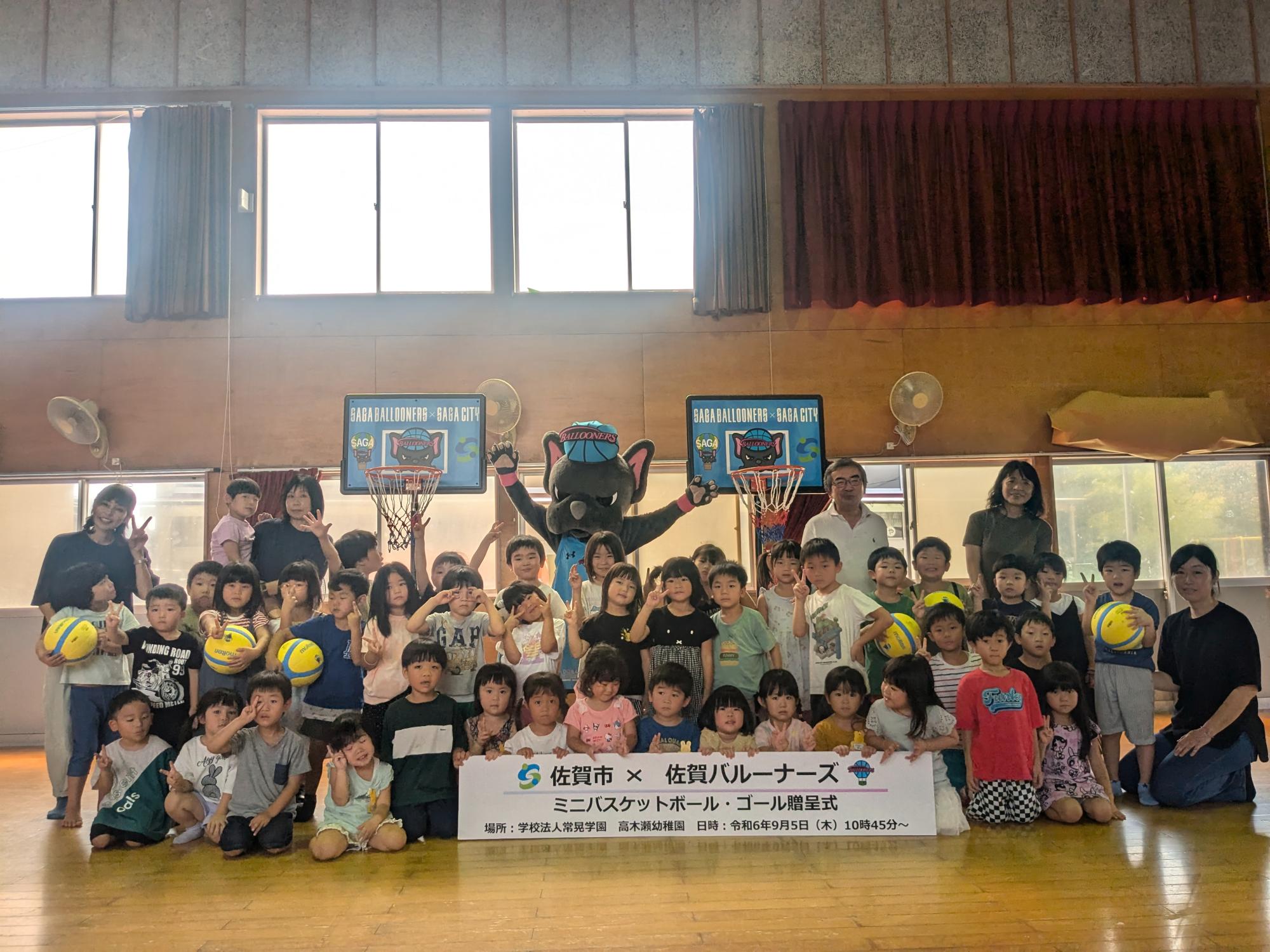 高木瀬幼稚園での贈呈式で、体育館に子供たちや職員の方が並び、その後ろにミニバスケットゴール2つ、手を広げたバルたんが立っている集合写真