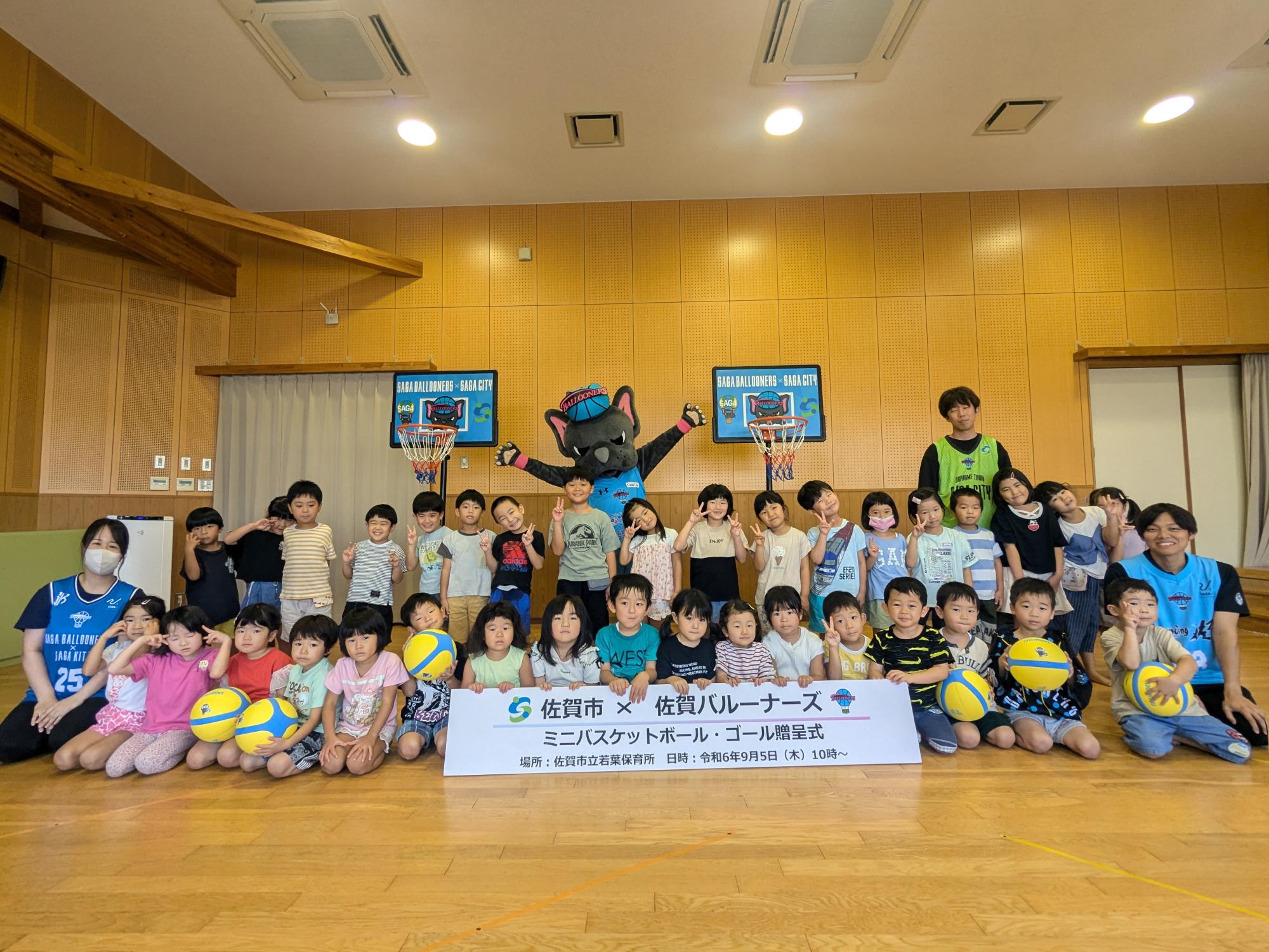 若葉保育所での贈呈式で、子供たちや佐賀バルーナーズの選手が並び、その後ろにミニバスケットゴール2つと両手を広げたパルたんが立っている集合写真