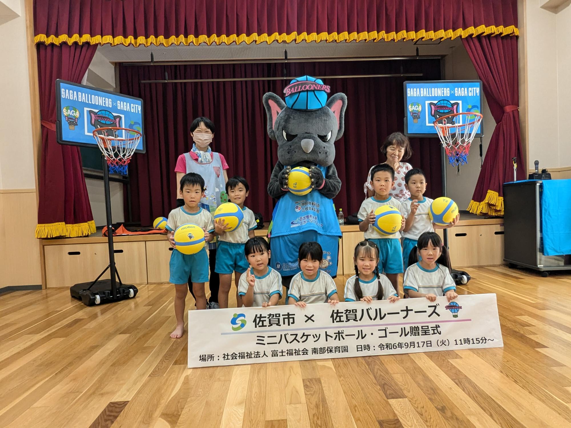 南部保育園での贈呈式で、前列に横断幕を持った4人、後列にボールを持った4人の子供たちが並び、後列中央にボールを持ったバルたんが立ち、両サイドにミニバスケットゴールが置かれている集合写真