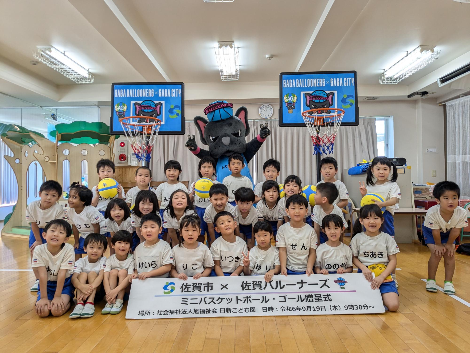 日新こども園での贈呈式で、子供たちが並んでいる後ろに手を広げたバルたんが立ち、その両サイドにミニバスケットゴールが置かれている集合写真