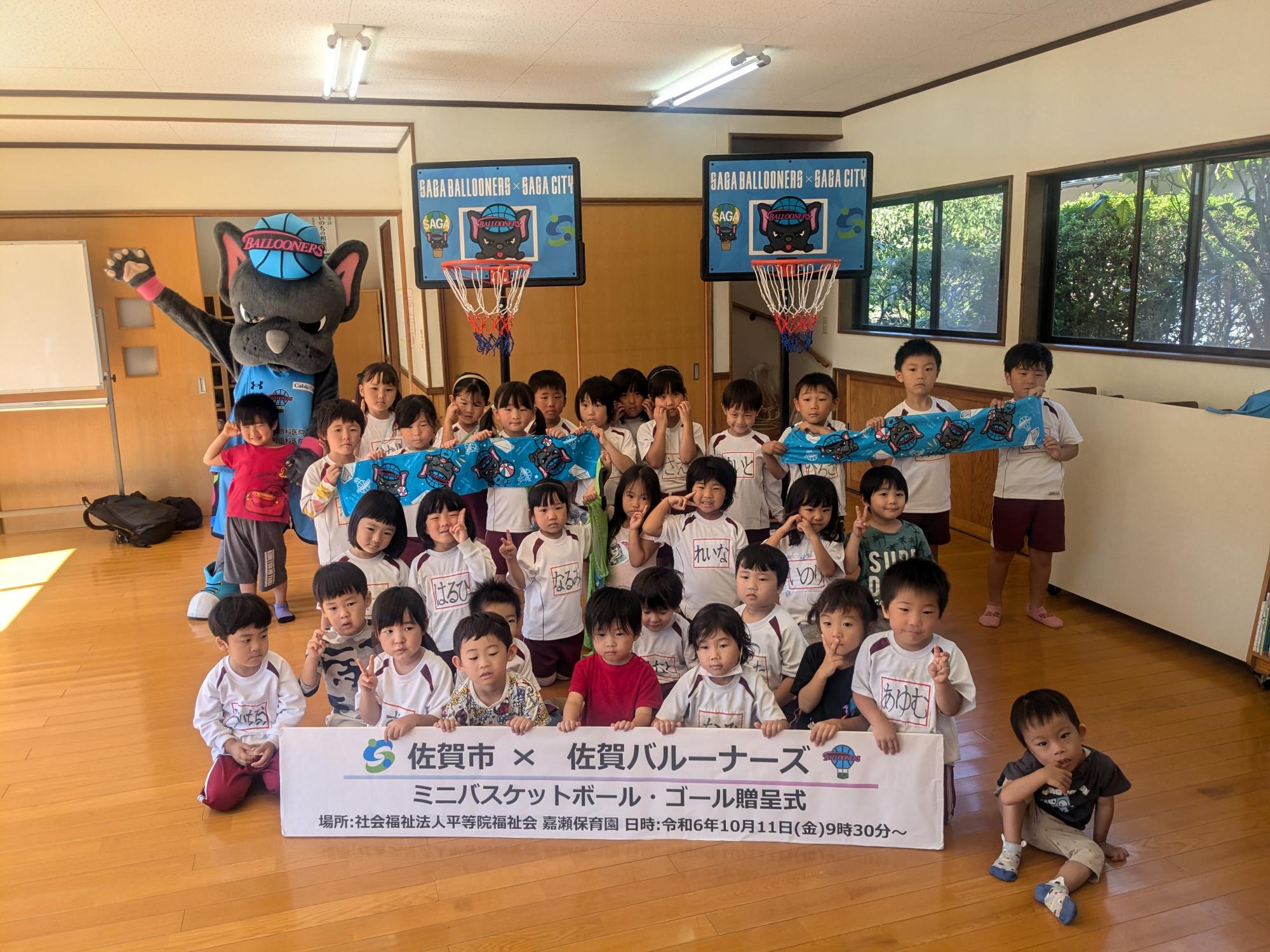 嘉瀬保育園での贈呈式で、30人ほどの子供たちが並んだ後ろに2つのミニバスケットゴールが置かれ、左側にバルたんが立っている集合写真