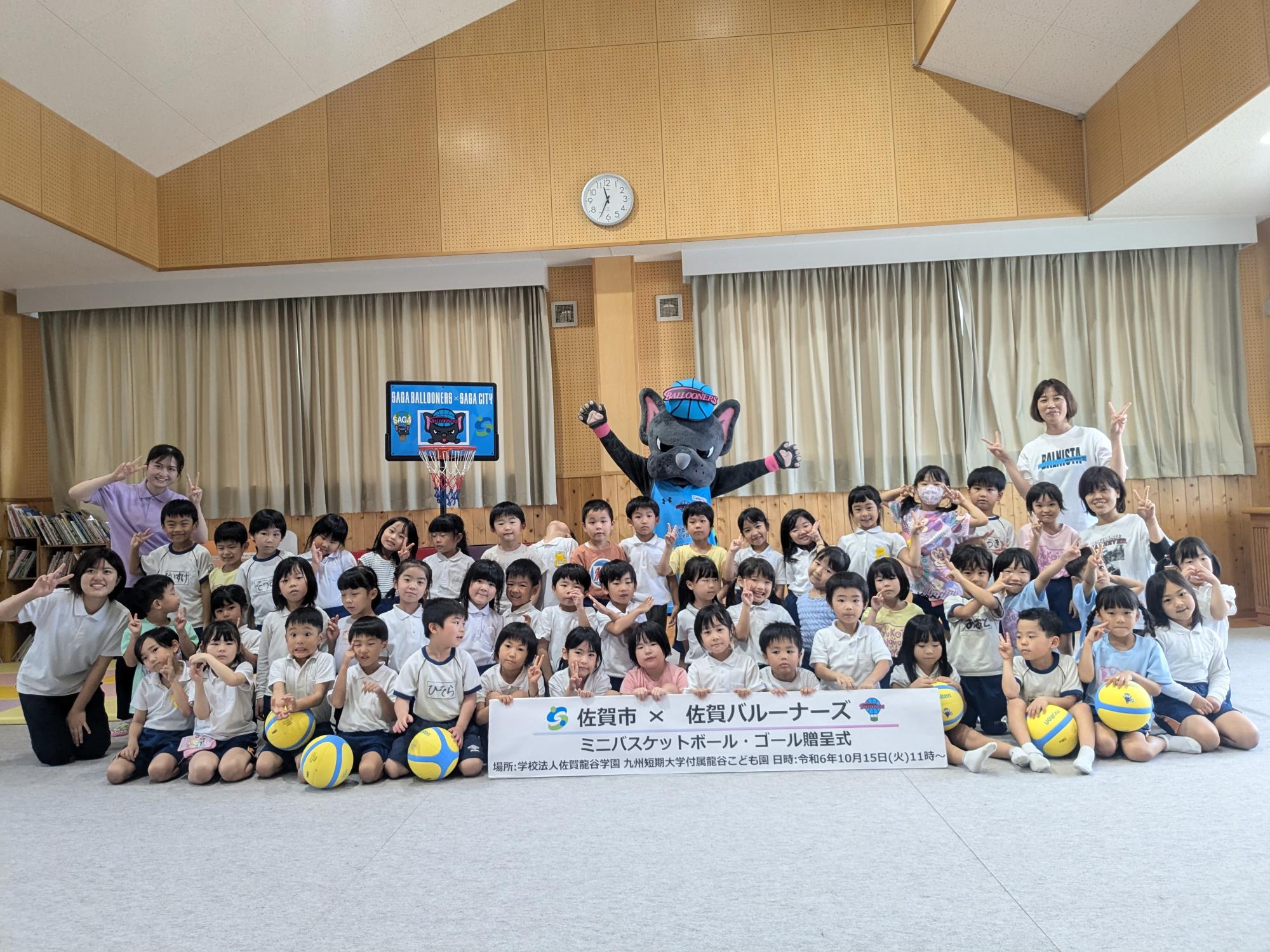 龍谷こども園での贈呈式で、3列に並んだ子供たちの後ろに両手を広げたバルたんが立ち、左側にミニバスケットゴールが置かれている集合写真