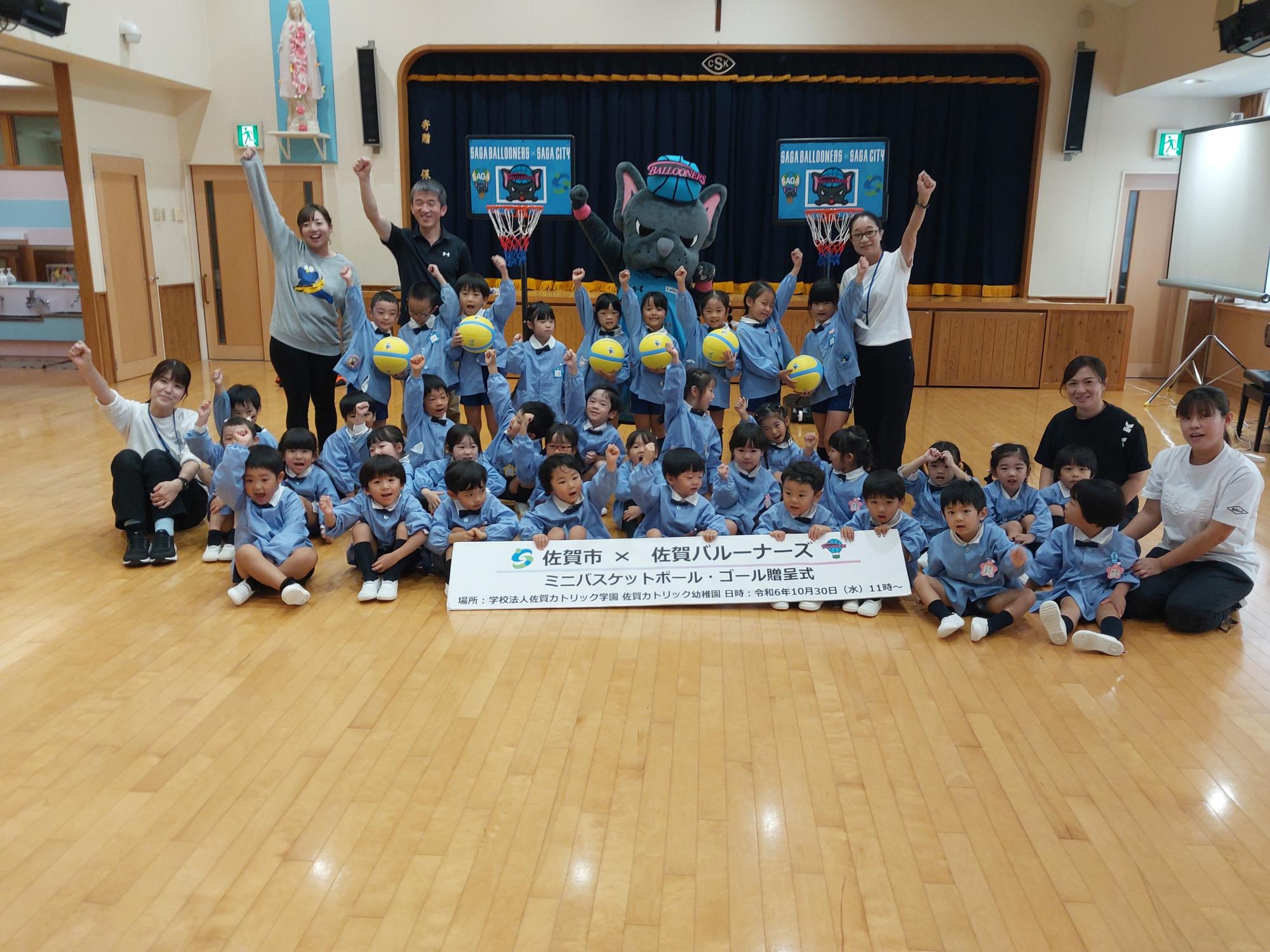 佐賀カトリック幼稚園での贈呈式で、集まって並んだ子供たちや職員の方が手をあげ、その後ろでバルたんも手をあげて立ち、両サイドにミニバスケットゴールが置かれている集合写真