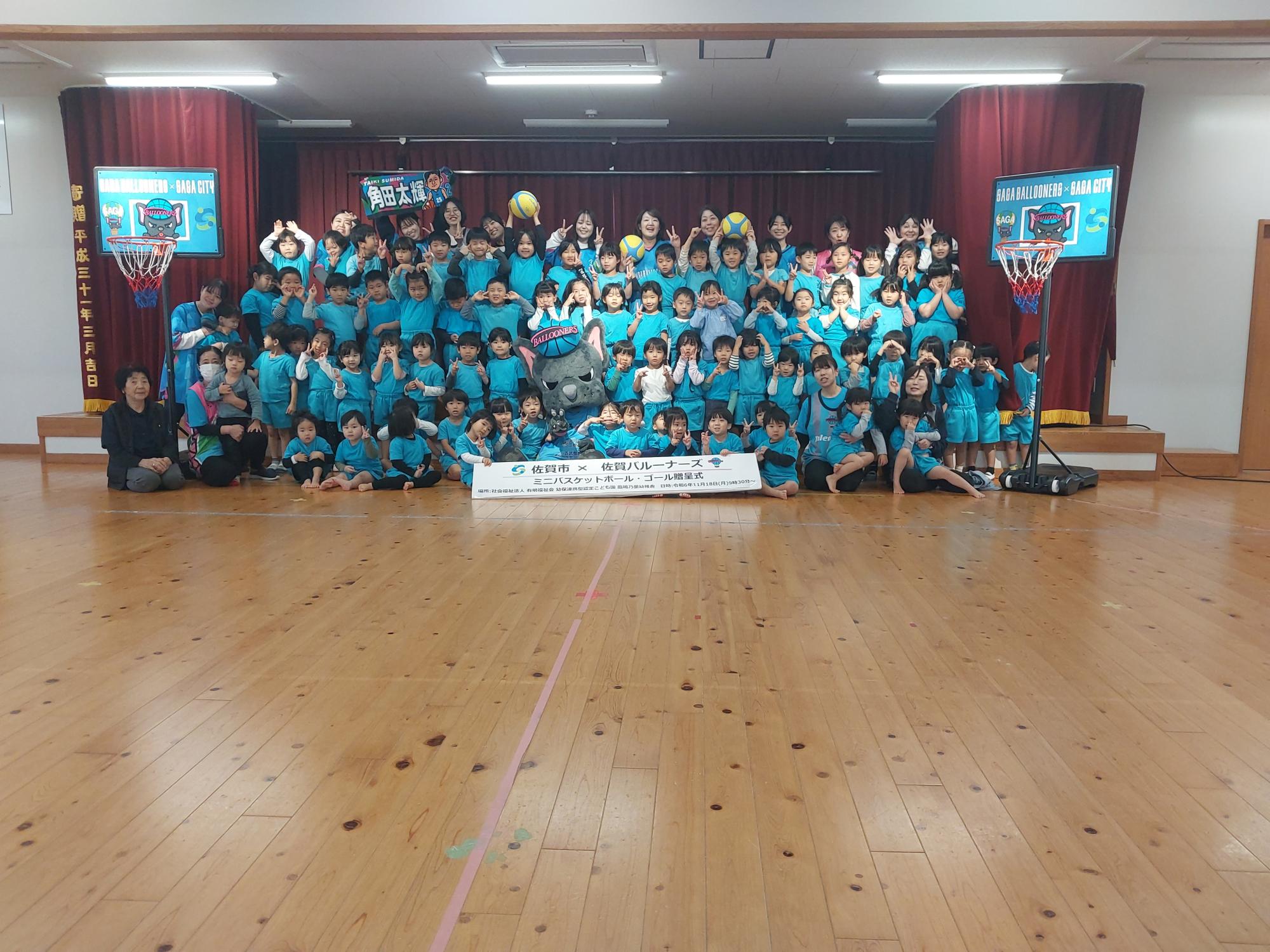 鳳鳴乃里幼稚園での贈呈式で、ステージ前列中央に座ってピースをするバルたんと周りにならんだ子供たちの集合写真