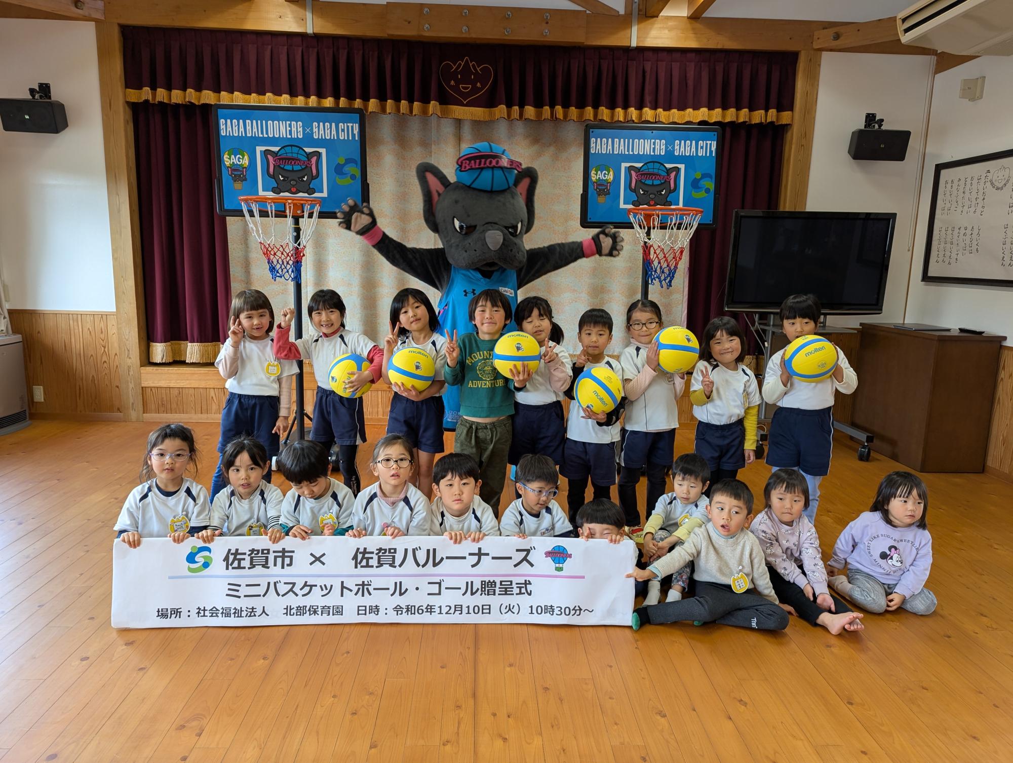 北部保育園での贈呈式で、2列に並んだ子供たちの後ろに両手を広げたバルたんがたち、両サイドにミニバスケットゴールが置かれている集合写真