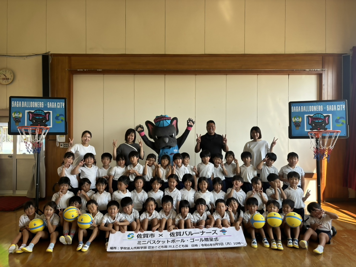 川上こども園での贈呈式で、3列に並んだ子供たちの後ろに佐賀バルーナーズの選手と両手を広げたバルたんが立ち、両サイドにミニバスケットゴールが置かれている集合写真