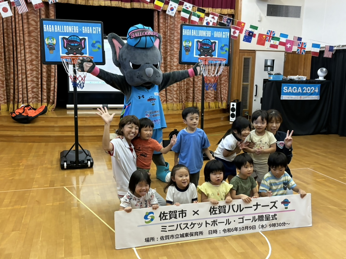 城東保育園での贈呈式で、2列に並んだ10人ほどの子供たちの後ろに手を広げたバルたんが立ち、その両サイドにミニバスケットゴールが置かれている集合