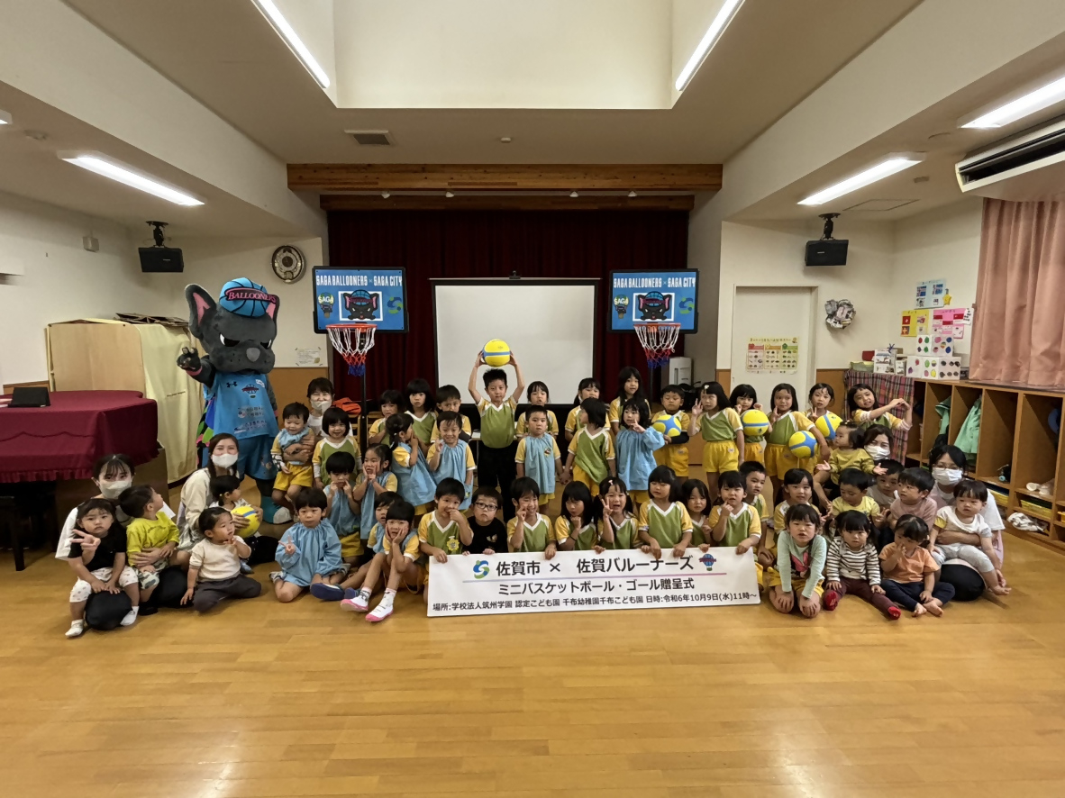 千布幼稚園での贈呈式で、4,50人ほどの子供たちが並んだ後ろに2つのミニバスケットゴールが置かれ、左側にバルたんが立っている集合写真