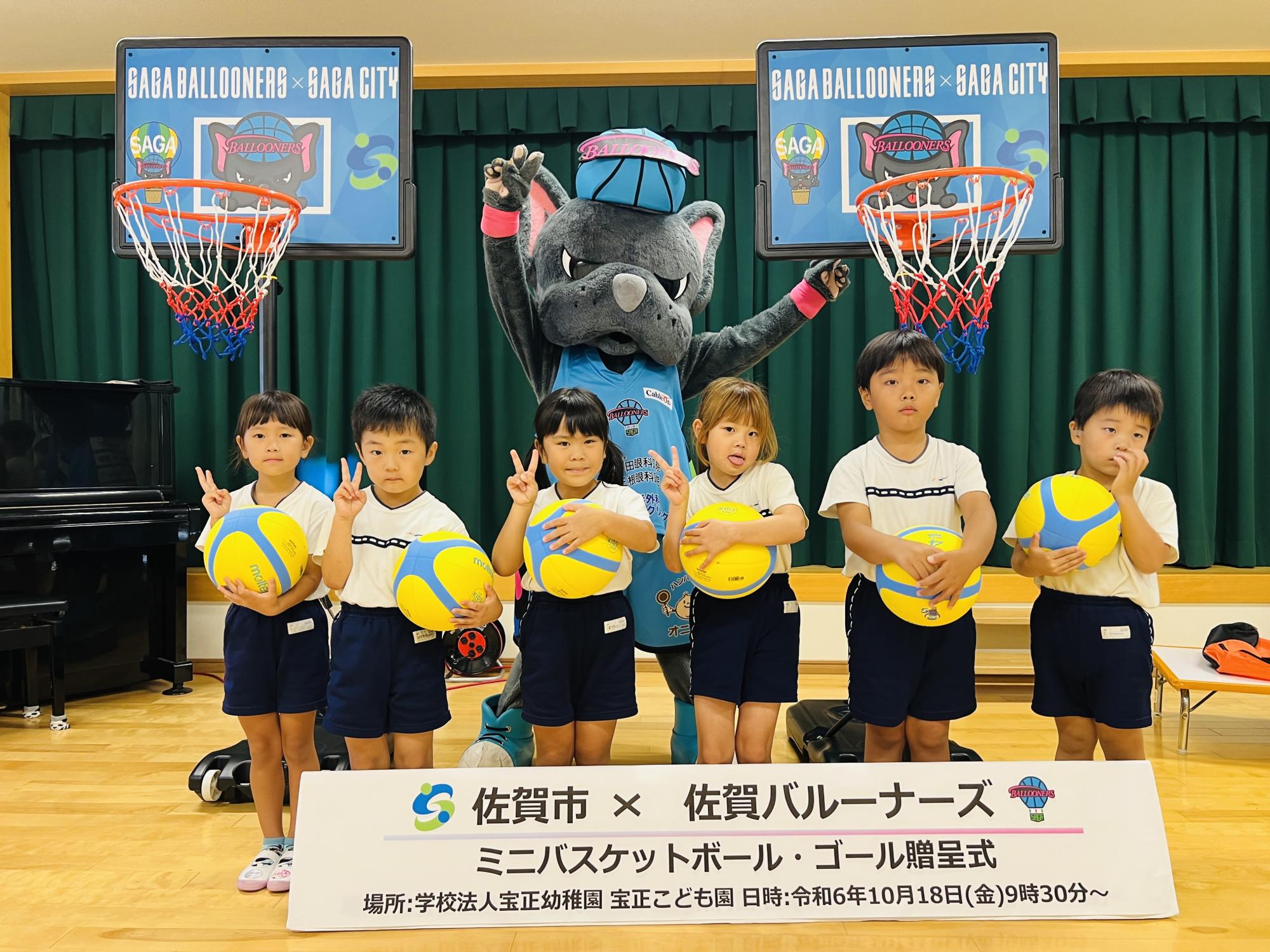 宝正こども園での贈呈式で、一列に並んでボールをもってピースをする6人の子供たちと、その後ろで両手を広げたバルたんが立ち、両サイドにミニバスケットゴールが置かれている集合写真