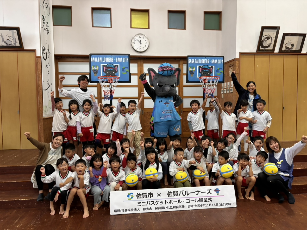 保育園ひなた村自然塾での贈呈式で、集まって並び手をあげる子供たちの中央に、手をあげたバルたんが立ち、両サイドにミニバスケットゴールが置かれている集合写真